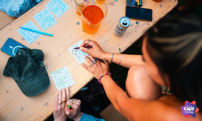 Spelers zitten aan een tafel buiten tijdens TAPT Festival met drankjes en bingo kaarten.