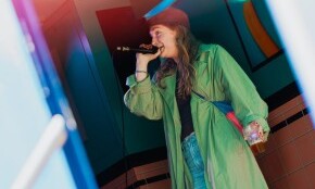 Persoon met microfoon en drankje, draagt groene jas op kleurrijke achtergrond.