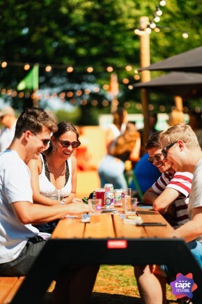 Vier mensen zitten aan een houten picknicktafel op een zonnig festival.