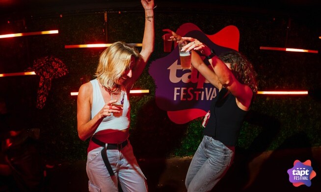 Twee personen dansen enthousiast op een feestje voor een bord met 'tapt festival'.