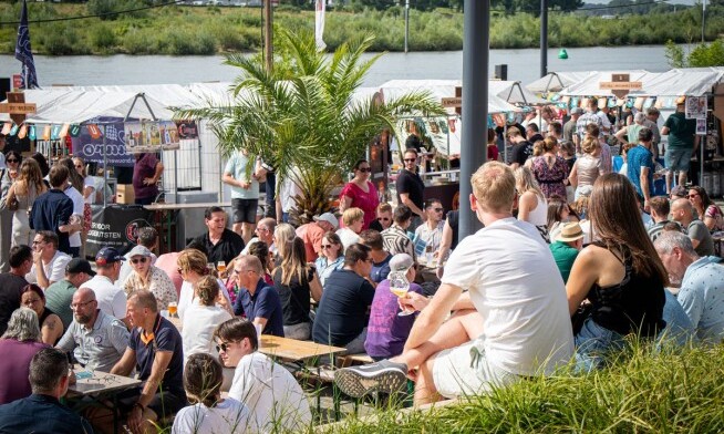 Groep mensen bij een bierfestival aan het water met kleurrijke kraampjes.