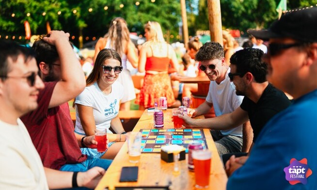 Groep mensen speelt een bordspel aan een houten tafel op een festival, zomerse sfeer.