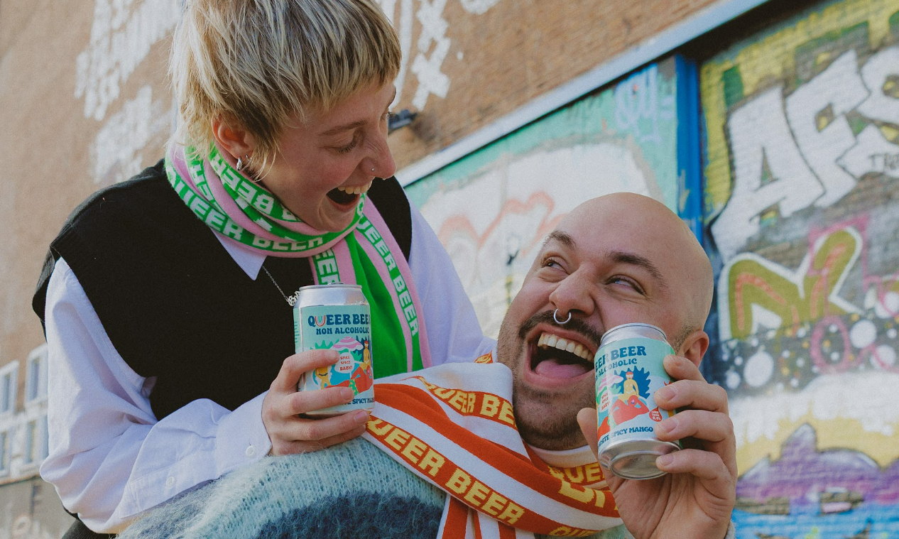 Twee mensen lachen samen terwijl ze Queer Beer 0.0 blikken vasthouden voor een met graffiti versierde muur.