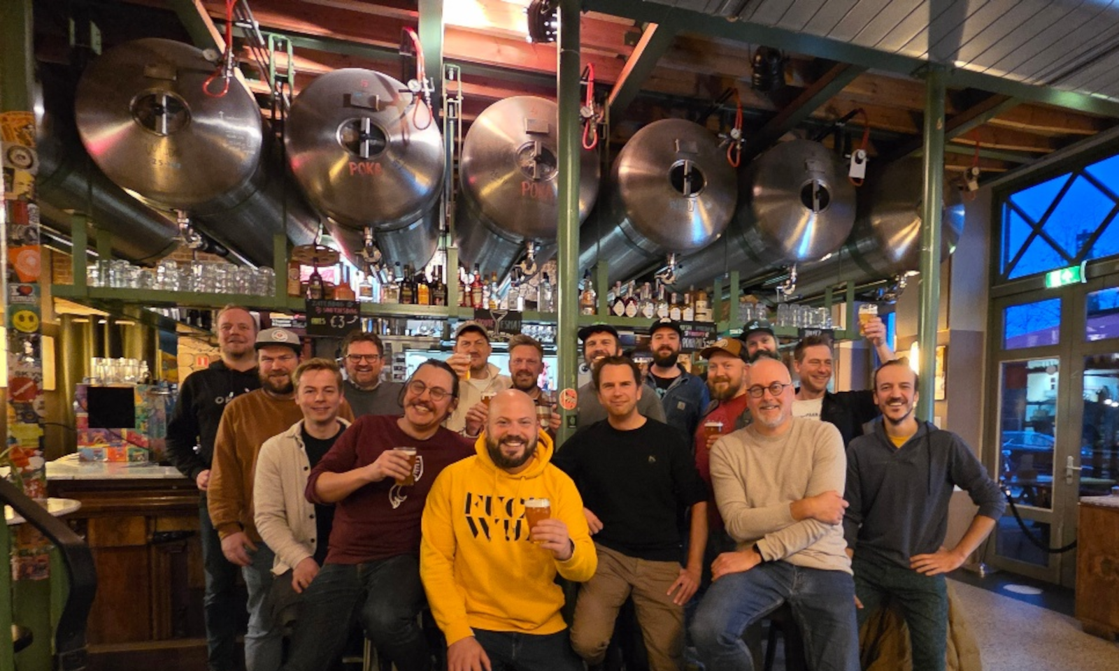 Brouwers betrokken bij CC in een brouwerij met grote tanks op de achtergrond, lachend met bier in de hand.