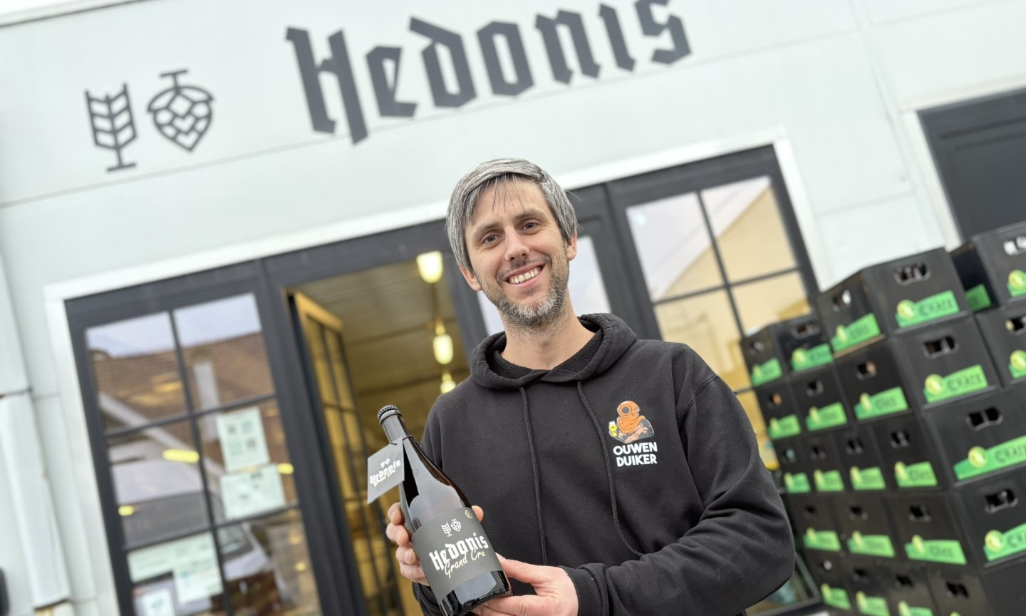 Man poseert met een fles Hedonis bier voor een brouwerij.