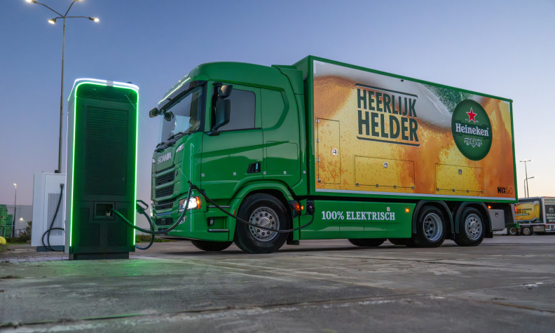 Een groene elektrische Heineken vrachtwagen wordt opgeladen bij een laadstation.
