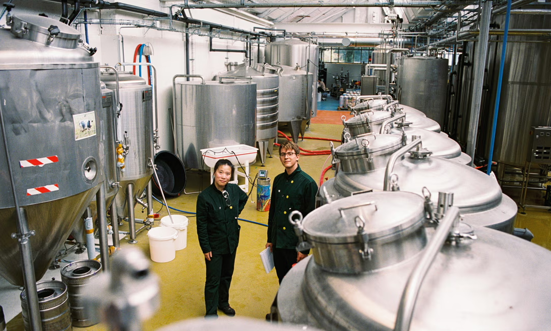 Twee mensen in groene werkkleding staan in een ruimte met grote stalen brouwketels.