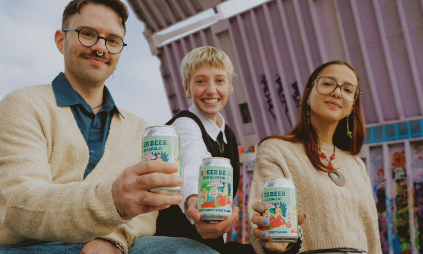 Drie personen houden Queer Beer blikjes omhoog, achtergrond met kleurrijke muur.