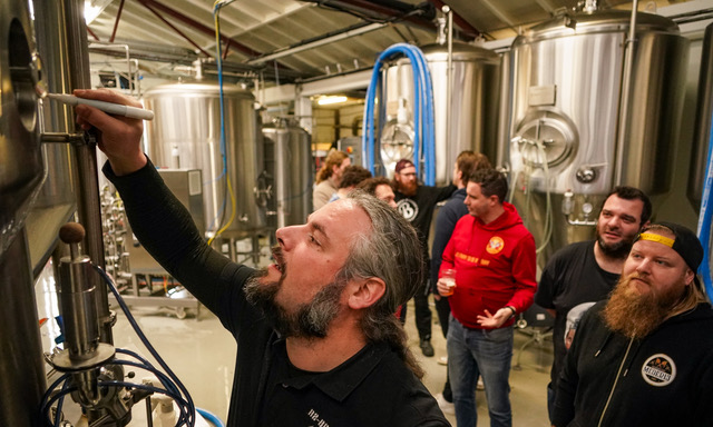 Brouwer schrijft op een roestvrijstalen tank in een brouwerij met andere mensen op de achtergrond.