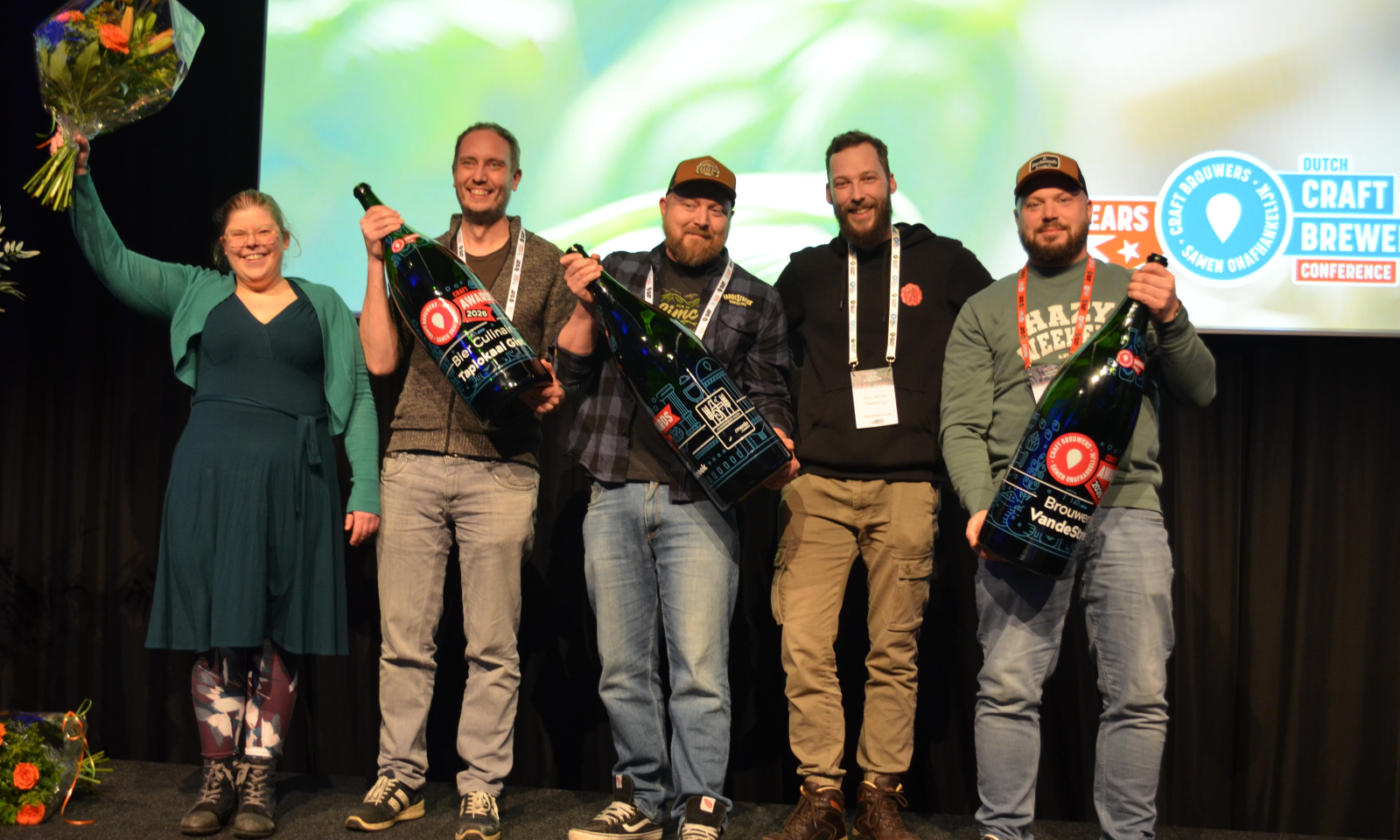 Groep mensen met grote flessen en bloemen poseren op een podium tijdens de CRAFT Awards 2026.