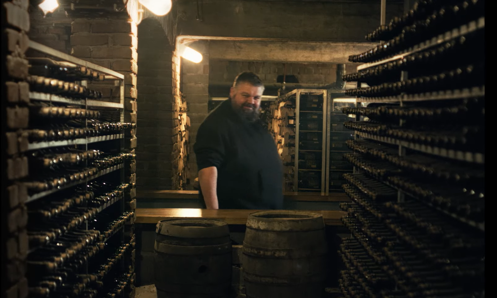 Man in een bierkelder met rijen flessen en houten vaten.