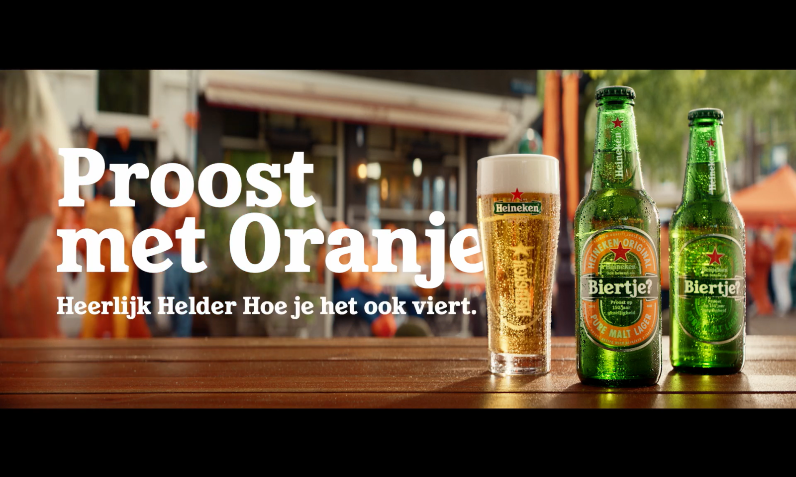 Twee Heineken bierflessen en een glas bier op een tafel met oranje decoratie op de achtergrond.