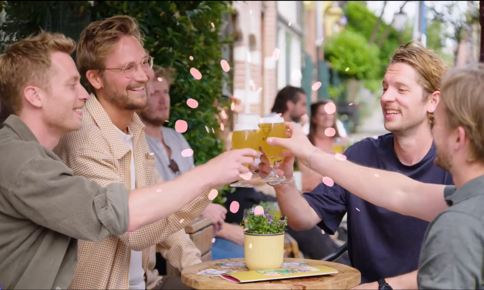 Groep vrienden proost met bier op zonnig terras met bloemblaadjes in de lucht.