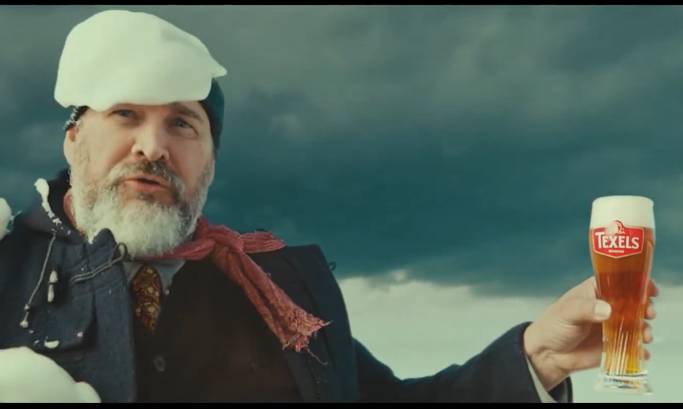 Man met baard en sneeuw op hoed, houdt een Texels bierglas tegen een bewolkte lucht.