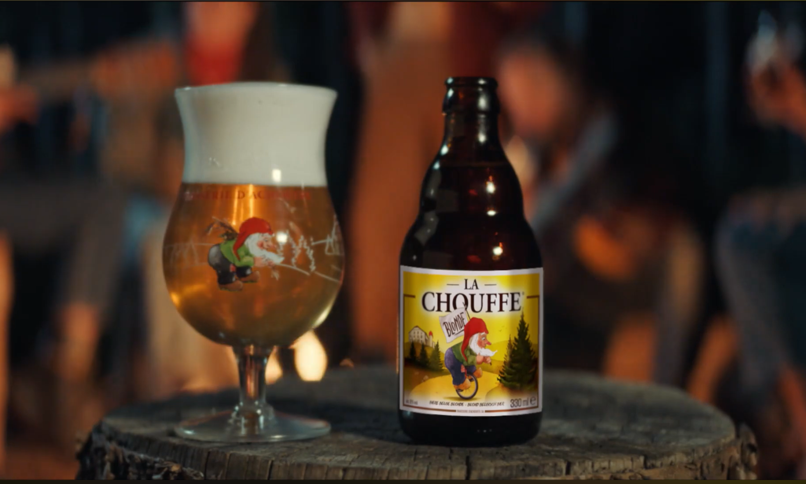 Bierglas en fles La Chouffe op een boomstronk met wazige achtergrond van mensen bij een kampvuur.
