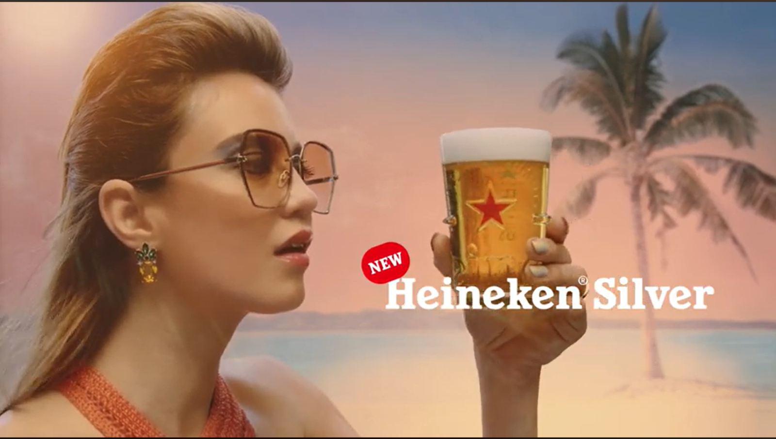 Vrouw met zonnebril houdt glas Heineken Silver bier vast op strand.