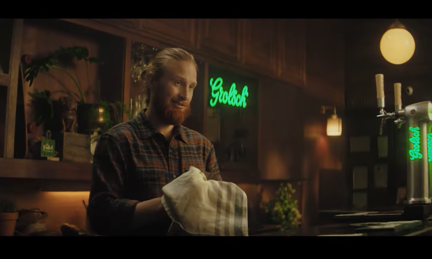 Barman met baard achter een bar met Grolsch neonlichten en tap.