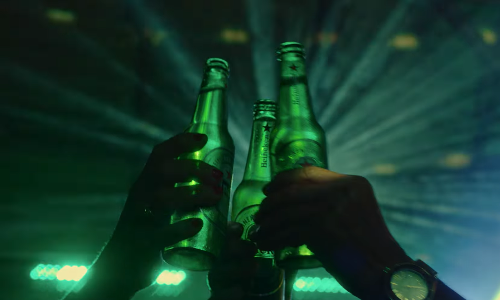 Drie handen proosten met groene bierflesjes tegen een achtergrond van feestverlichting.