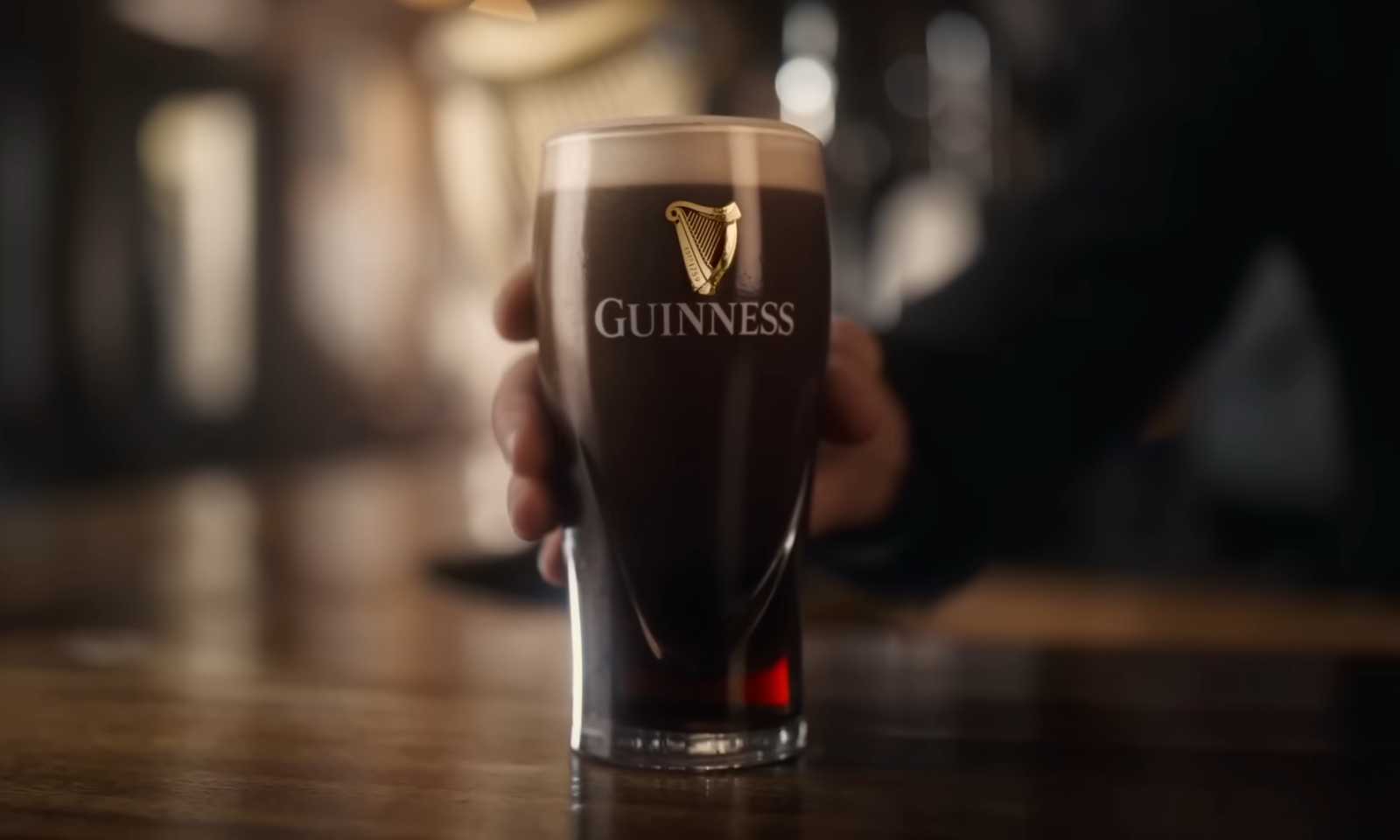 Een glas Guinness-bier wordt vastgehouden door een hand in een gezellige baromgeving.