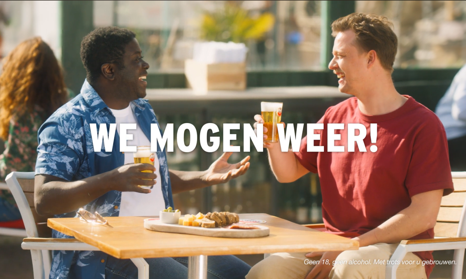Twee lachende mannen heffen hun bierglazen op een zonnig terras, met tekst 