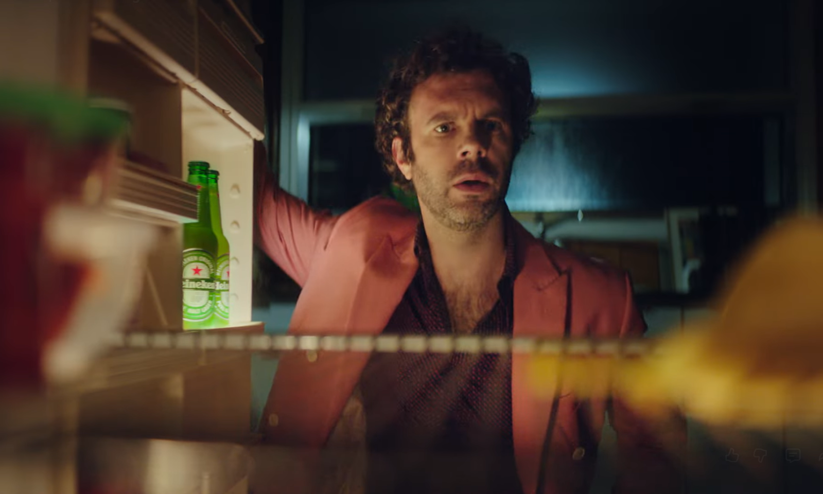 Man in roze jasje staat voor een open koelkast met flessen bier.