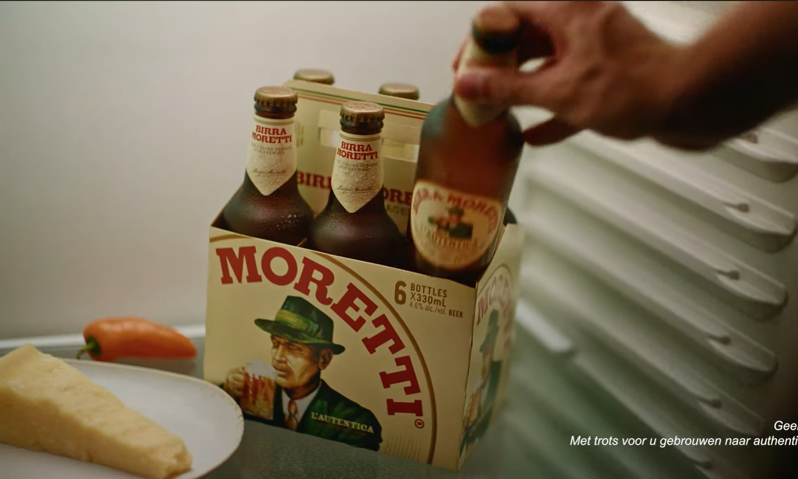 Hand pakt fles Birra Moretti uit kartonnen verpakking in koelkast naast kaas en peper.