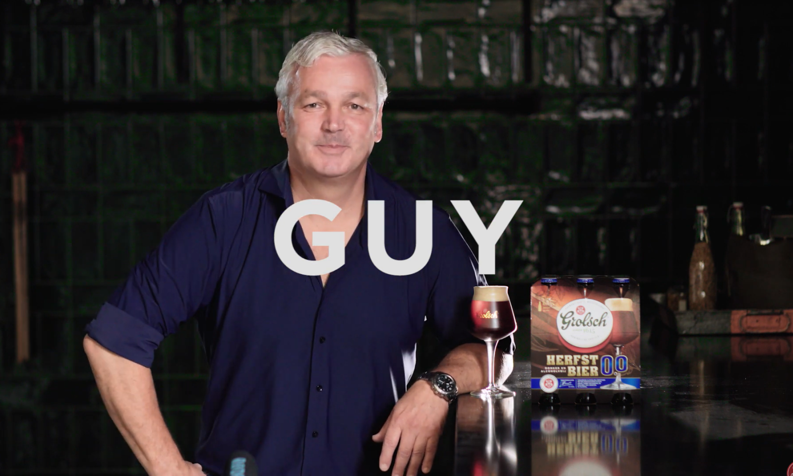 Man in blauw overhemd naast glas en verpakking Grolsch Herfstbier 0.0% op donkere achtergrond.