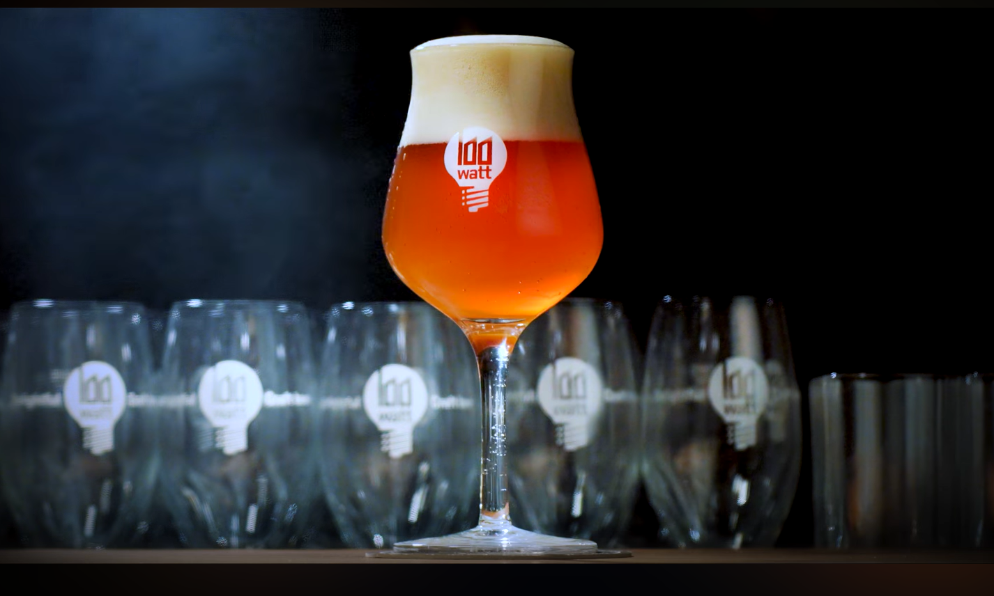 Een glas ambachtelijk bier met het logo van 100 Watt ernaast meer lege glazen.
