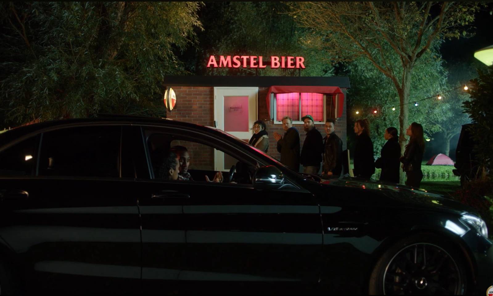 Mensen staan in de rij voor een kiosk met een Amstel Bier witte neonreclame erboven.