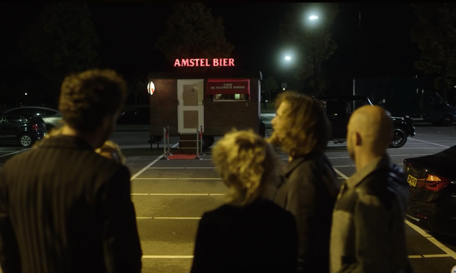 Een groep mensen kijkt naar een verlichte kiosk van Amstel Bier op een parkeerplaats.