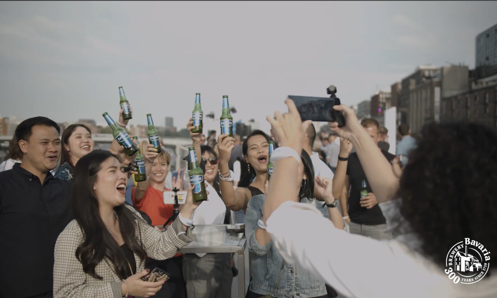 Groep mensen heft Bavaria bierflesjes in de lucht tijdens een evenement.