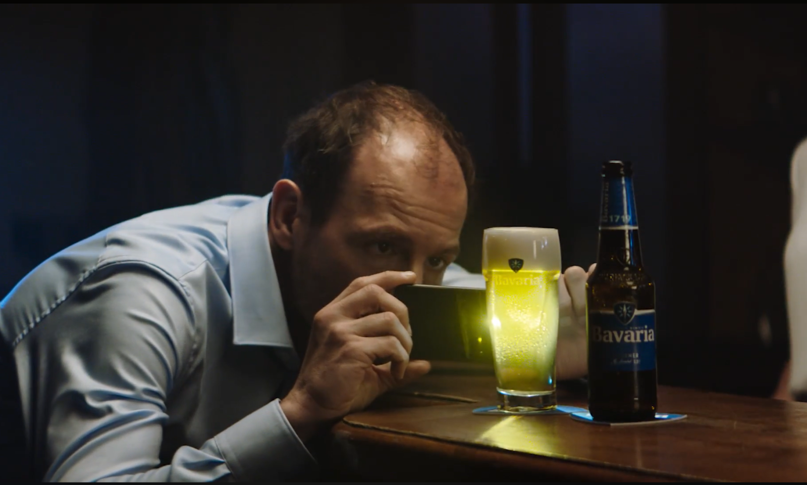 Man fotografeert een glas en fles Bavaria bier op een houten bar met zijn smartphone.