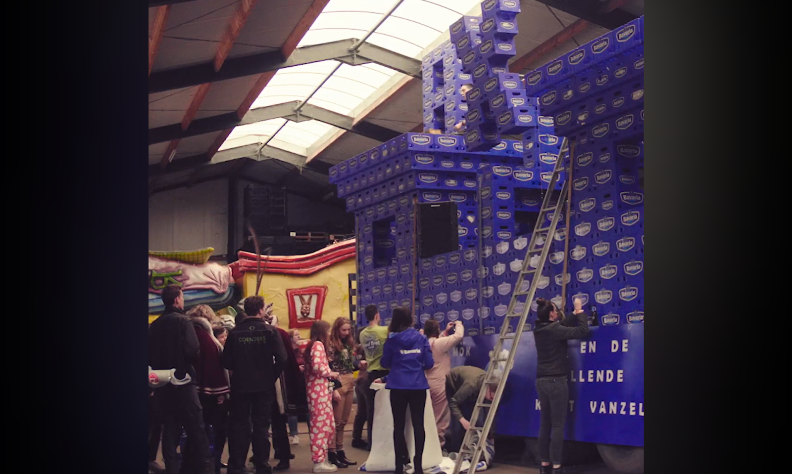 Personen maken een carnavalswagen van blauwe Bavaria kratten in een loods.