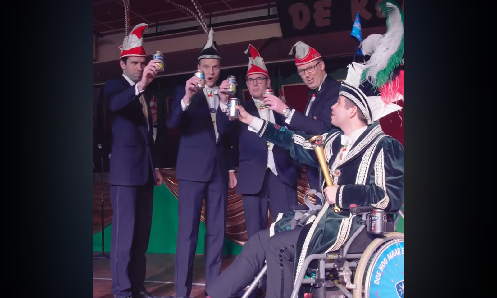 Vijf mannen in carnavalspakken proosten met bier, man in rolstoel vooraan.