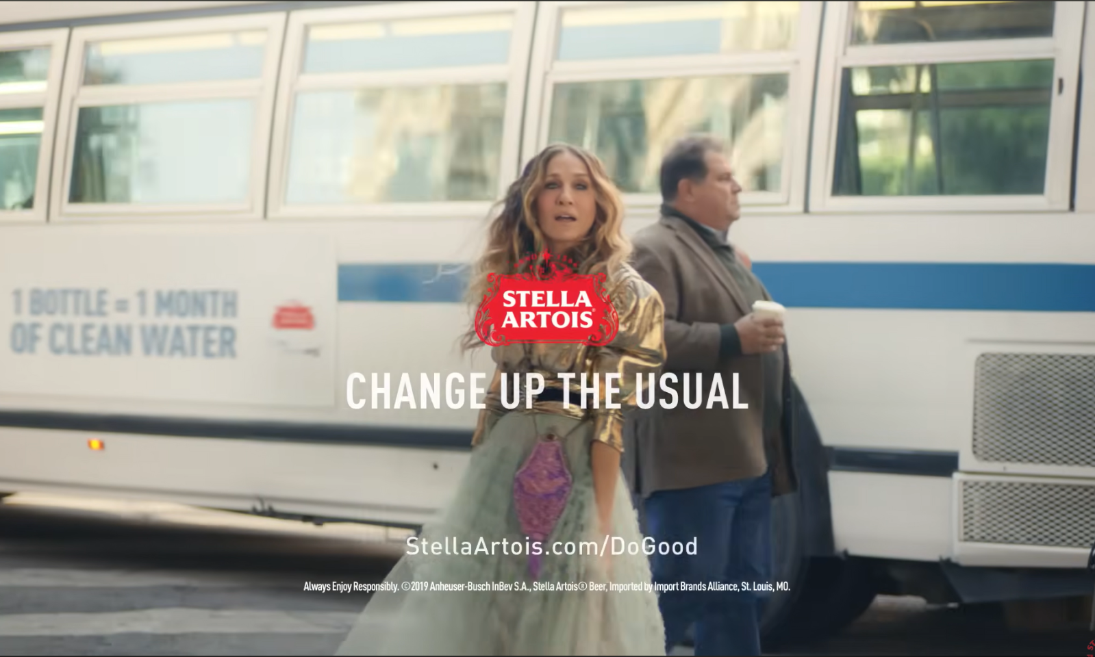 Vrouw in gouden jurk met Stella Artois logo voor een langsrijdende bus in stad.