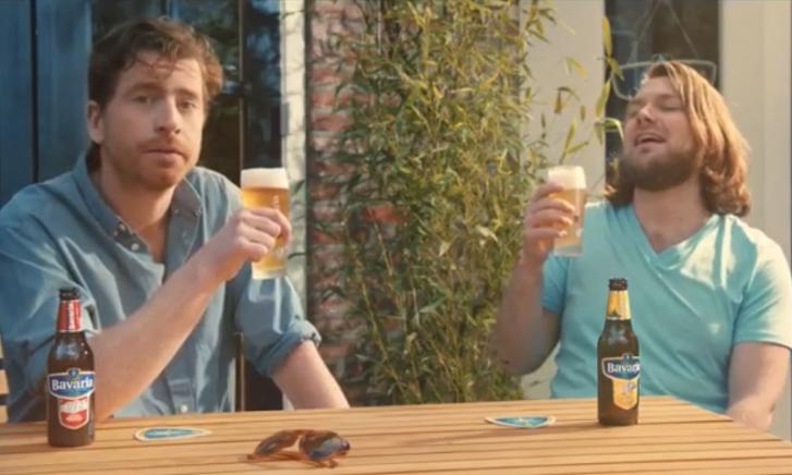 Man met halflang haar lacht en wijst, zittend aan tafel met Bavaria fles.