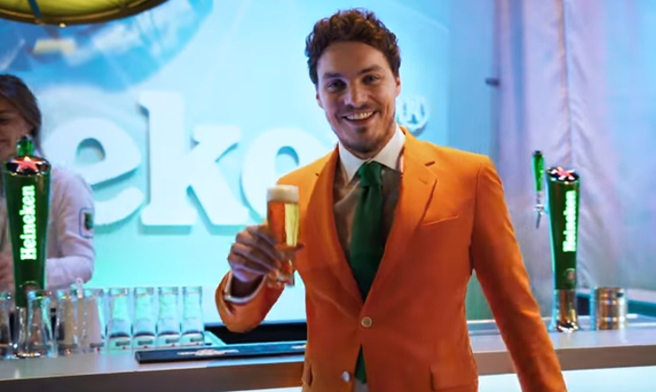 Een man in een oranje pak met groene stropdas staat voor het Heineken-logo.