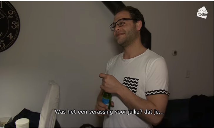 Groep mensen proost met groene bierflessen in een woonkamer met klok aan de muur.