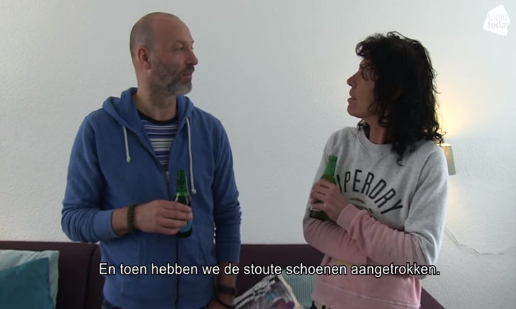 Twee lachende mensen die proosten met flesjes drank in een woonkamer.