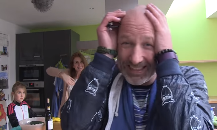 Gezin in keuken met man die fles inspecteert en vrouw die lacht naast een kind.