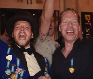 Man in blauwe jas met medailles en hoge hoed, tekst onderin beeld.