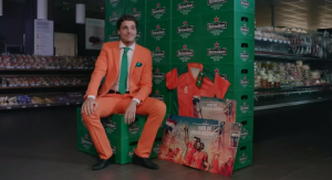 Man in Oranje Heineken sambashirt danst in supermarkt gangpad naast koelschappen.