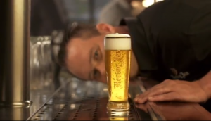 Drie mannen aan de bar, één drinkt een glas bier met schuimkraag.